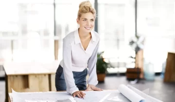 Smiling female contractor with blueprint at desk looking away in office. Horizontal shot.
