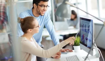 Two colleagues having business discussion in modern office, woman analyst showing successful financial and marketing statistics from screen to her male co-worker, sitting at desk with laptop computer