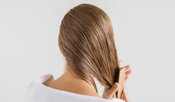 Woman in the white towel with comb brushing her wet blonde hair after shower on the gray background. Cares about a healthy and clean hair. Beauty salon concept.